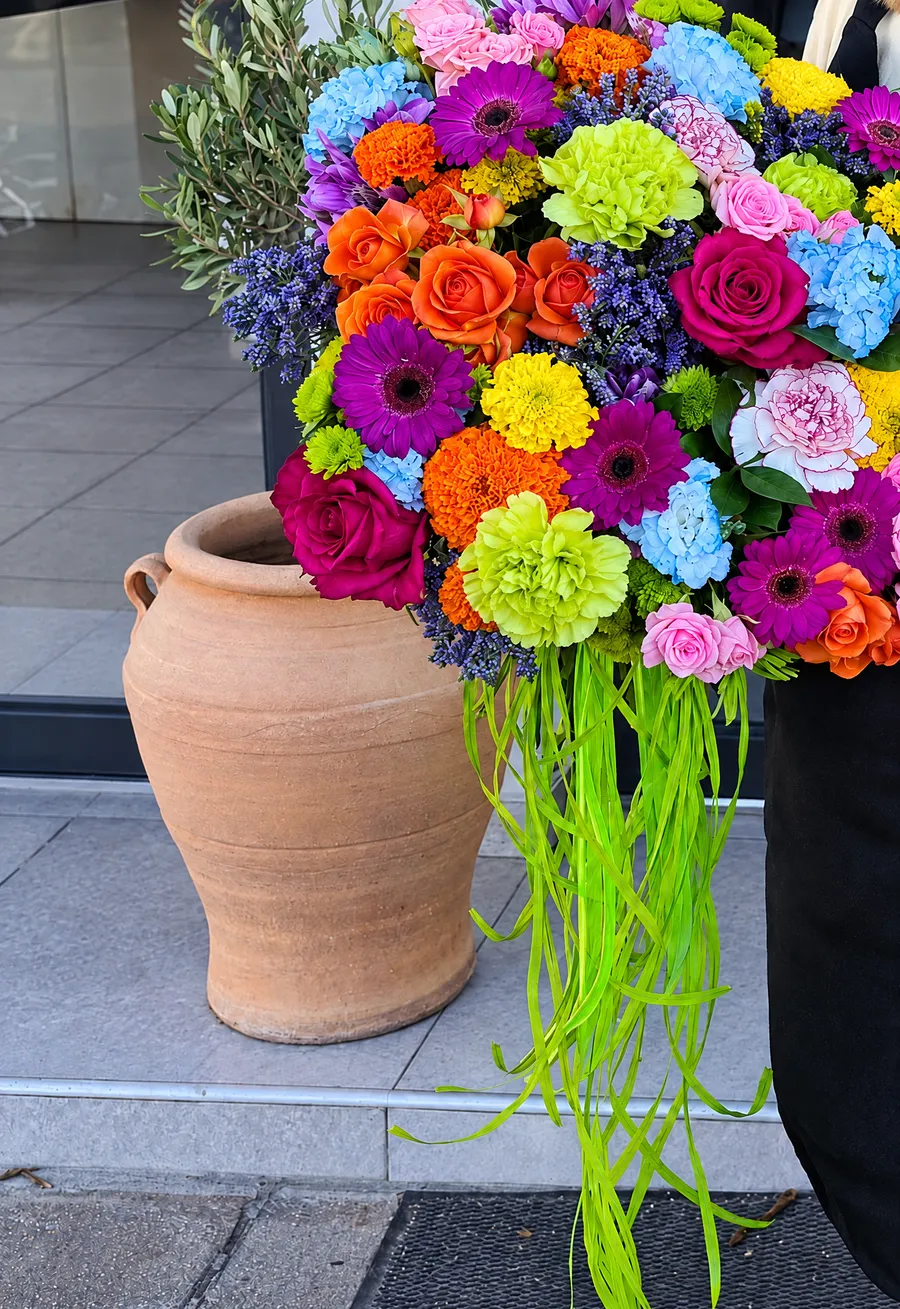 A colourful bouquet with long green ribbons beside a clay pithari at the shop door